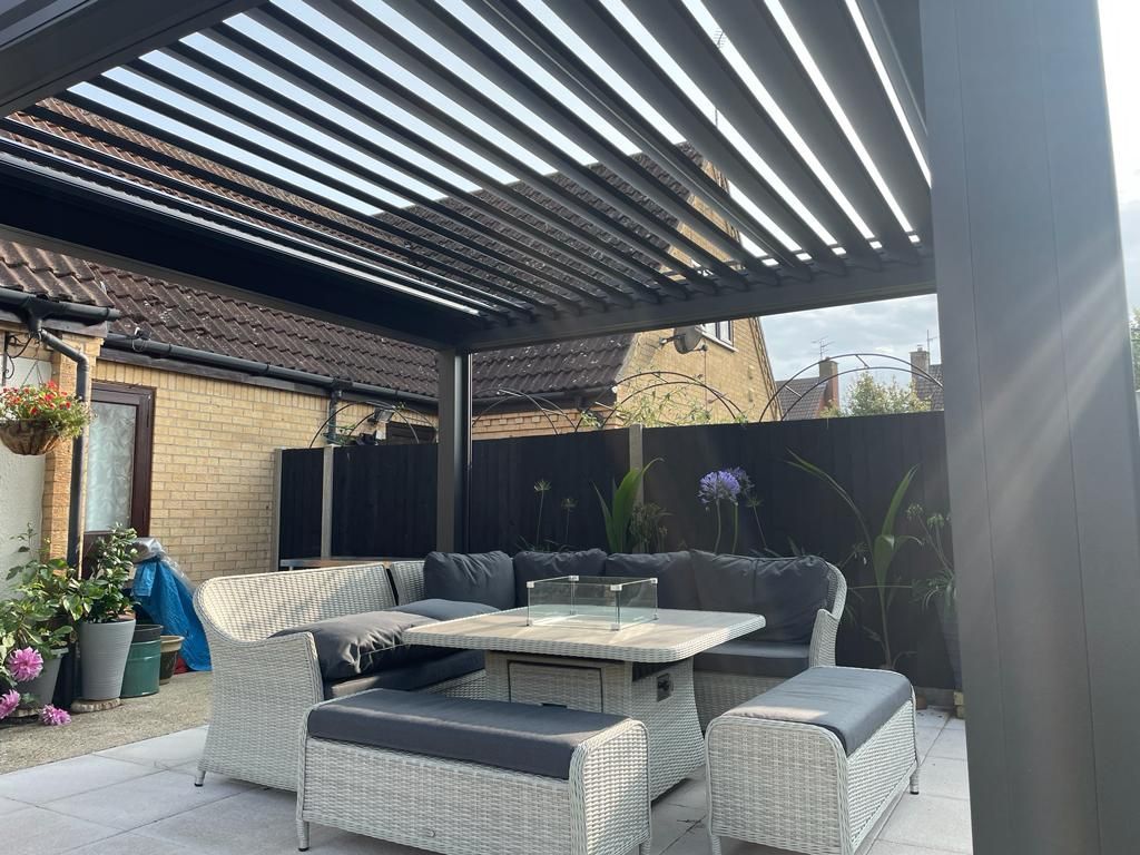 Patio with a pergola, gray furniture, and a stone table. The sun shines through the adjustable roof slats.