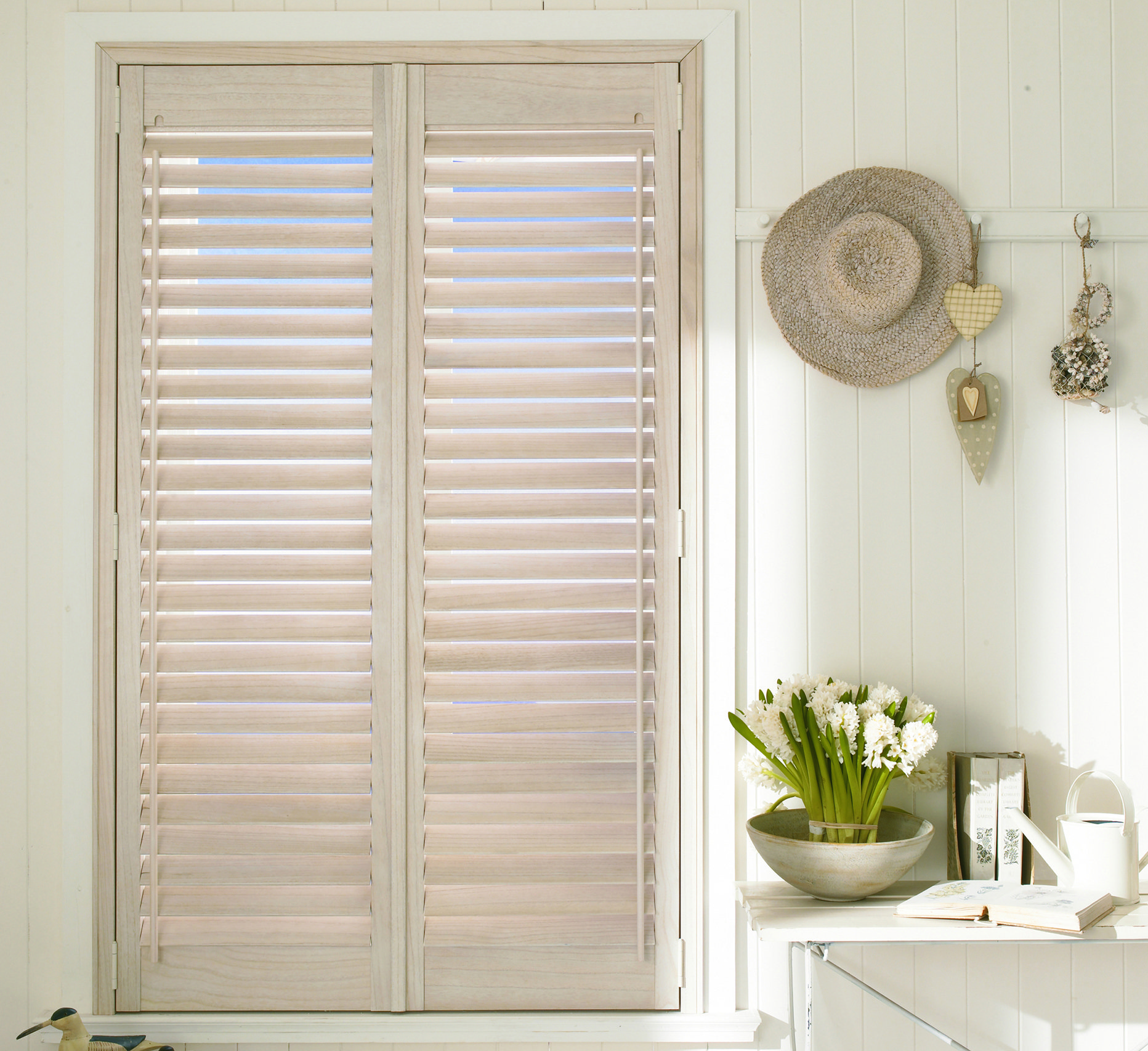 White-washed wooden shutters on a window; a hat, flowers, and a small desk decorate the windowsill.