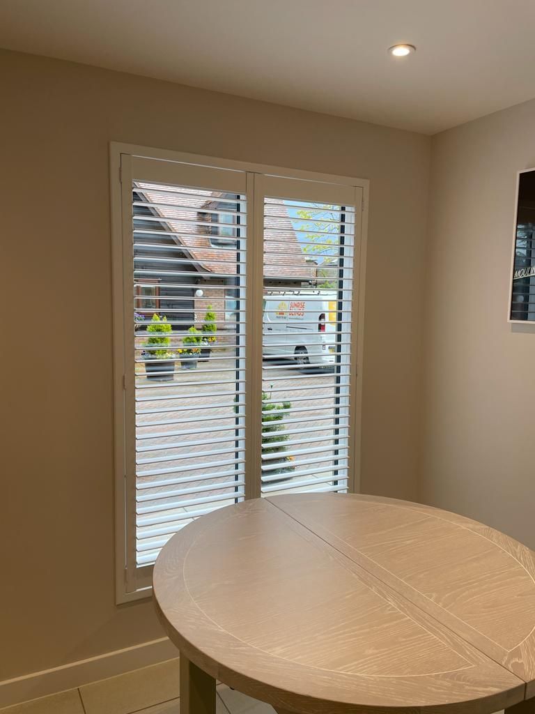 A round wooden table in front of a window with white shutters. Building visible outside.