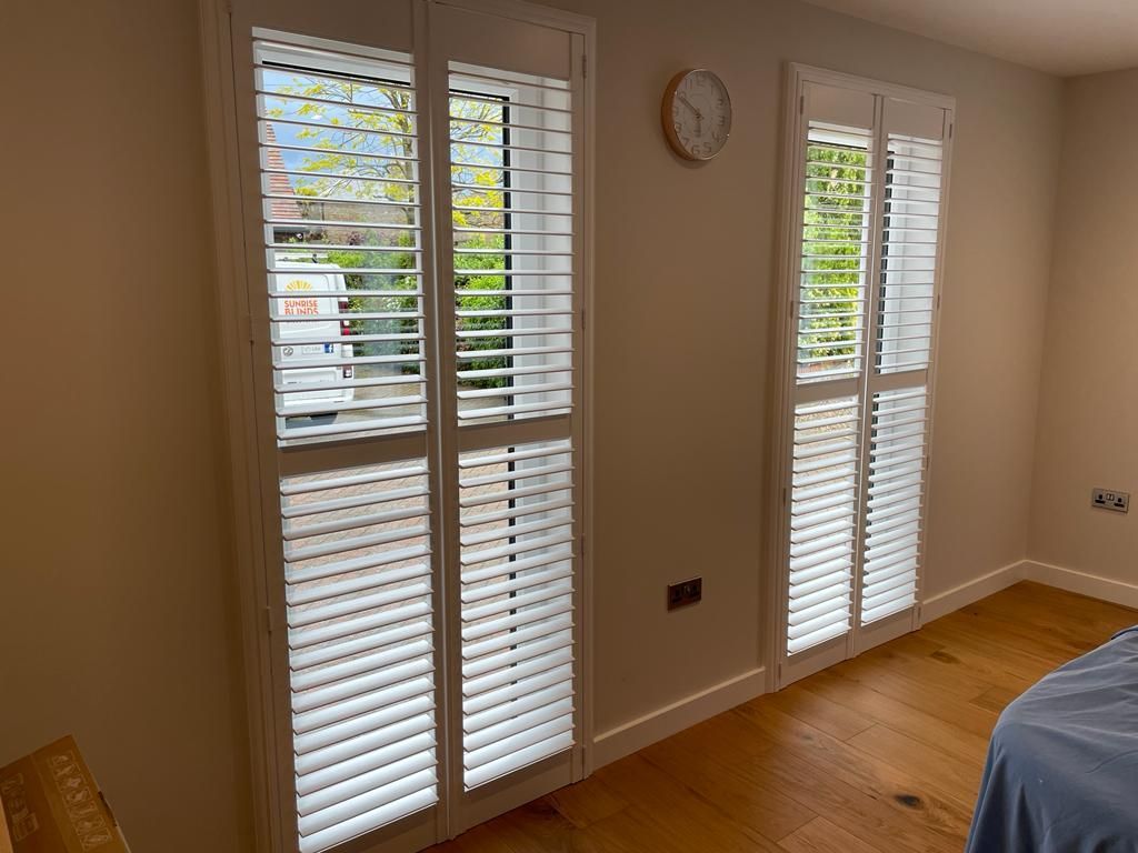 White interior shutters on two windows, with light wood flooring. A clock hangs on the wall.