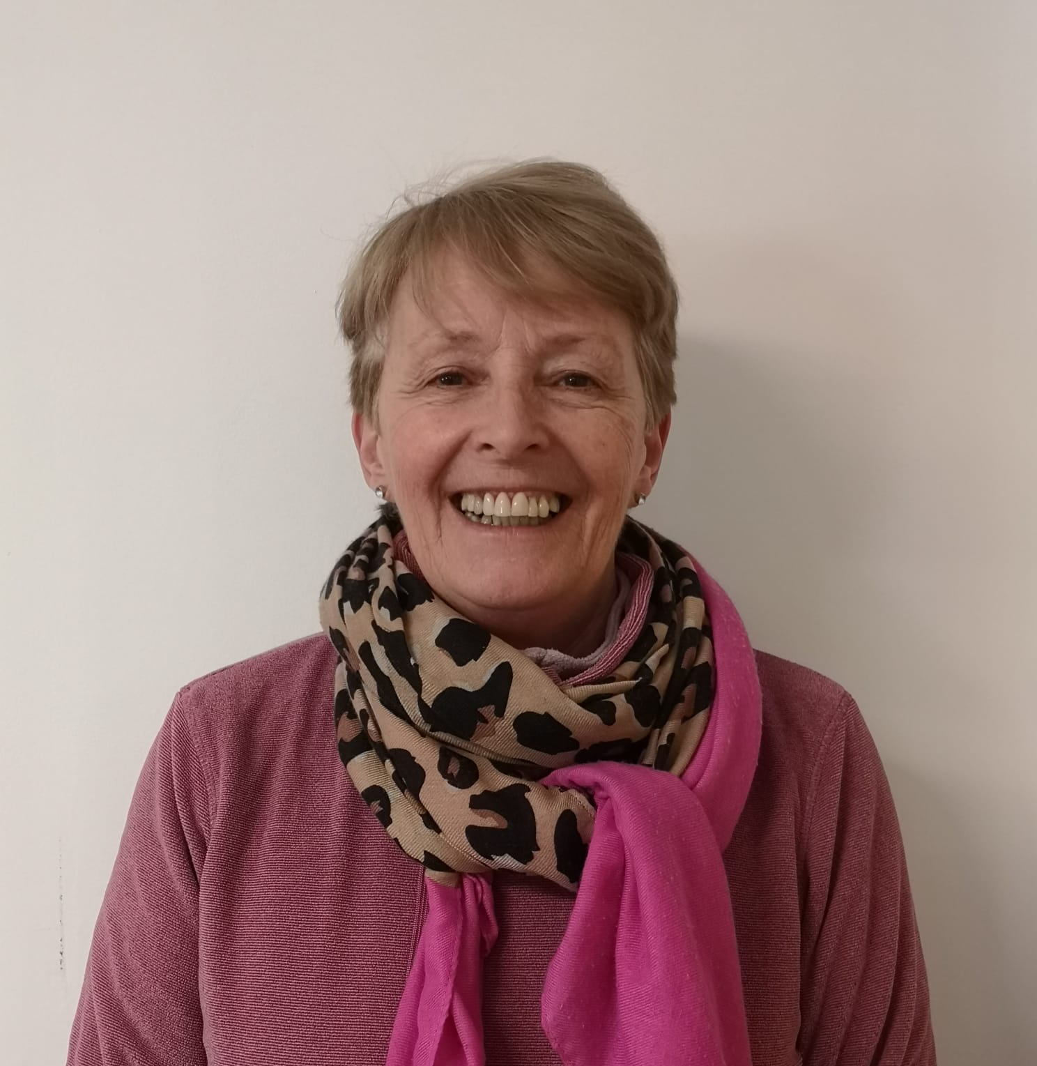 Woman with short hair, smiling, wearing a pink shirt and scarf with a tan and black animal print.