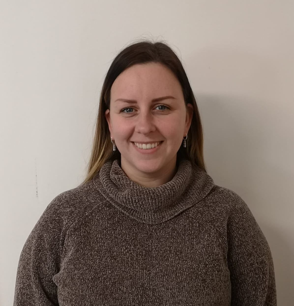 Woman smiling, wearing a brown turtleneck, against a plain white wall.