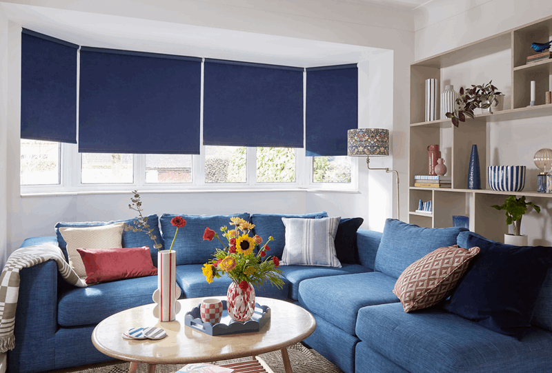 Blue sectional sofa in a bright living room with dark blue roller shades. Wooden shelves and round coffee table.