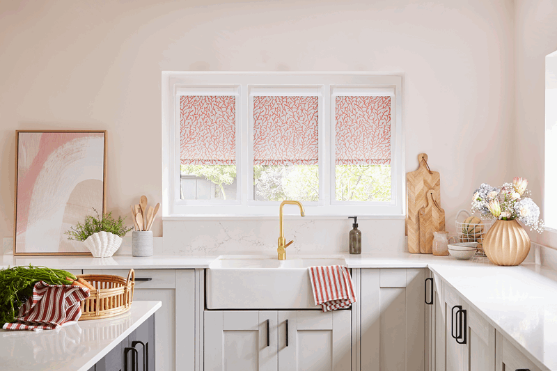 Kitchen with pale pink walls, white cabinets, and a window with floral blinds; gold faucet.