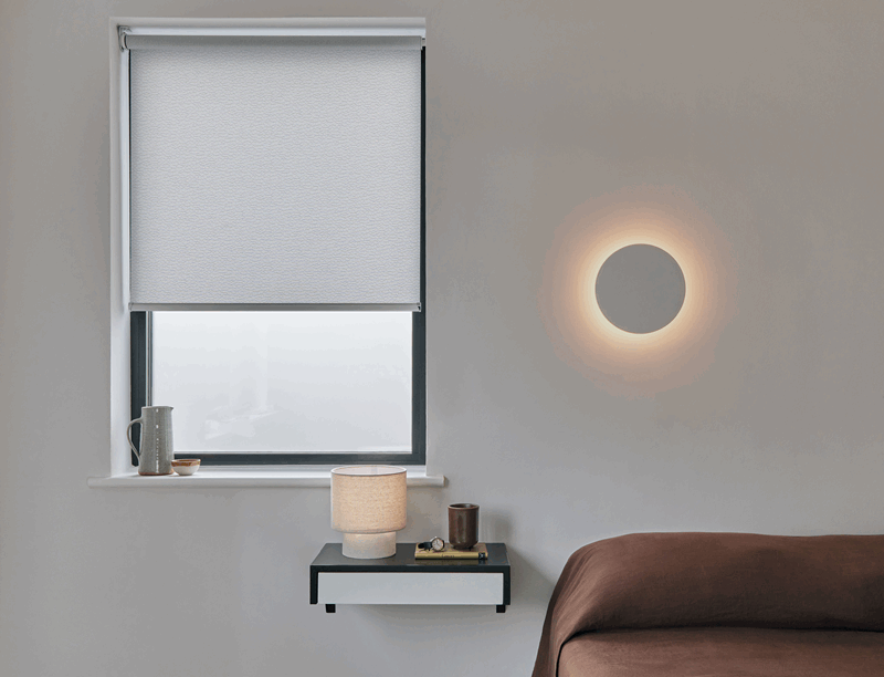 Bedroom scene with a window, white roller shade, bedside table, and a circular wall light.