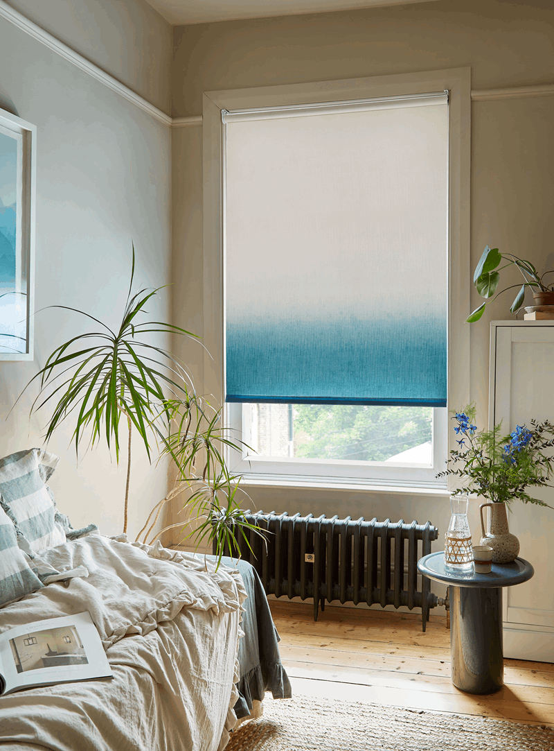 Bedroom with ombre blue-white window shade, plants, bed, small table, and radiator.