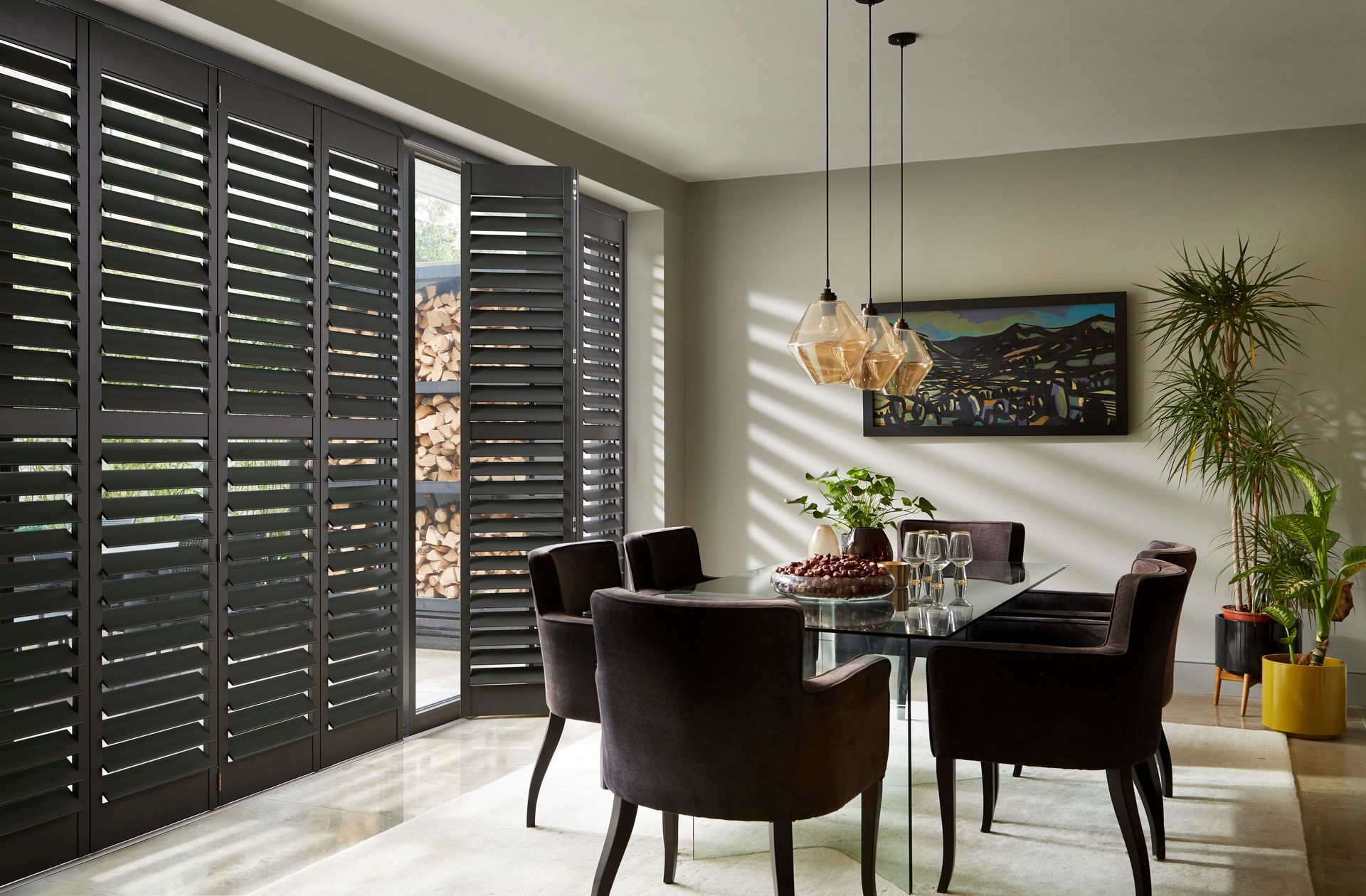Dining room with dark shutters, glass table, velvet chairs, and hanging pendant lights.