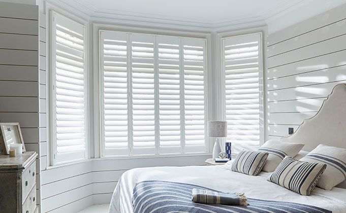 Bedroom with white shutters on bay windows, bed with blue and white pillows, white paneled walls.