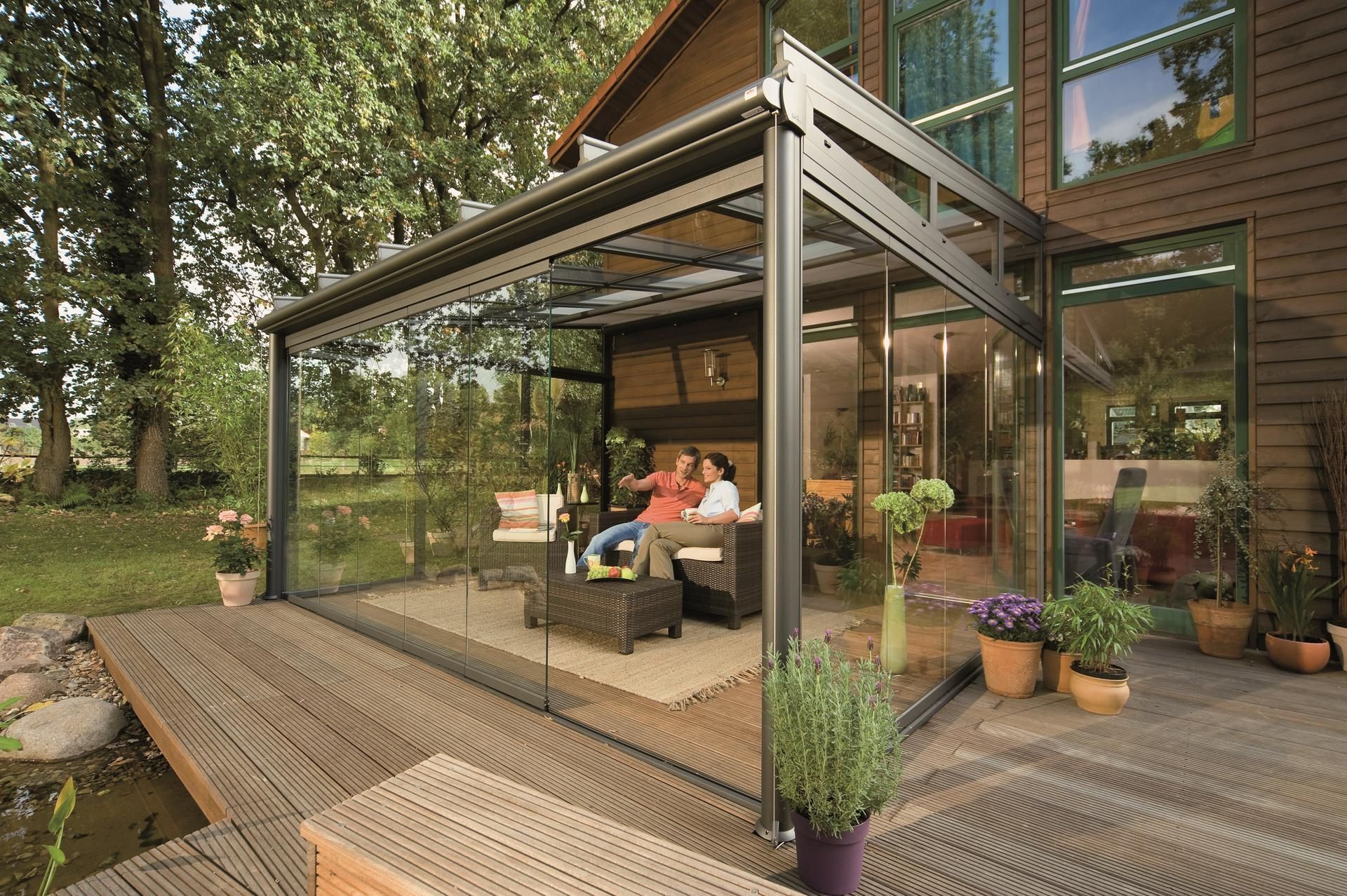 Glass-enclosed sunroom attached to a house. Two people sit on a sofa, plants nearby.