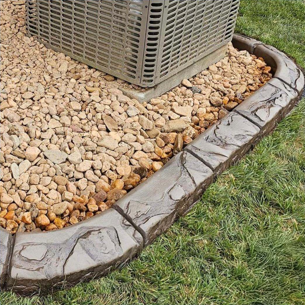 A textured concrete border separates a gravel bed containing an AC unit from a green grass lawn.