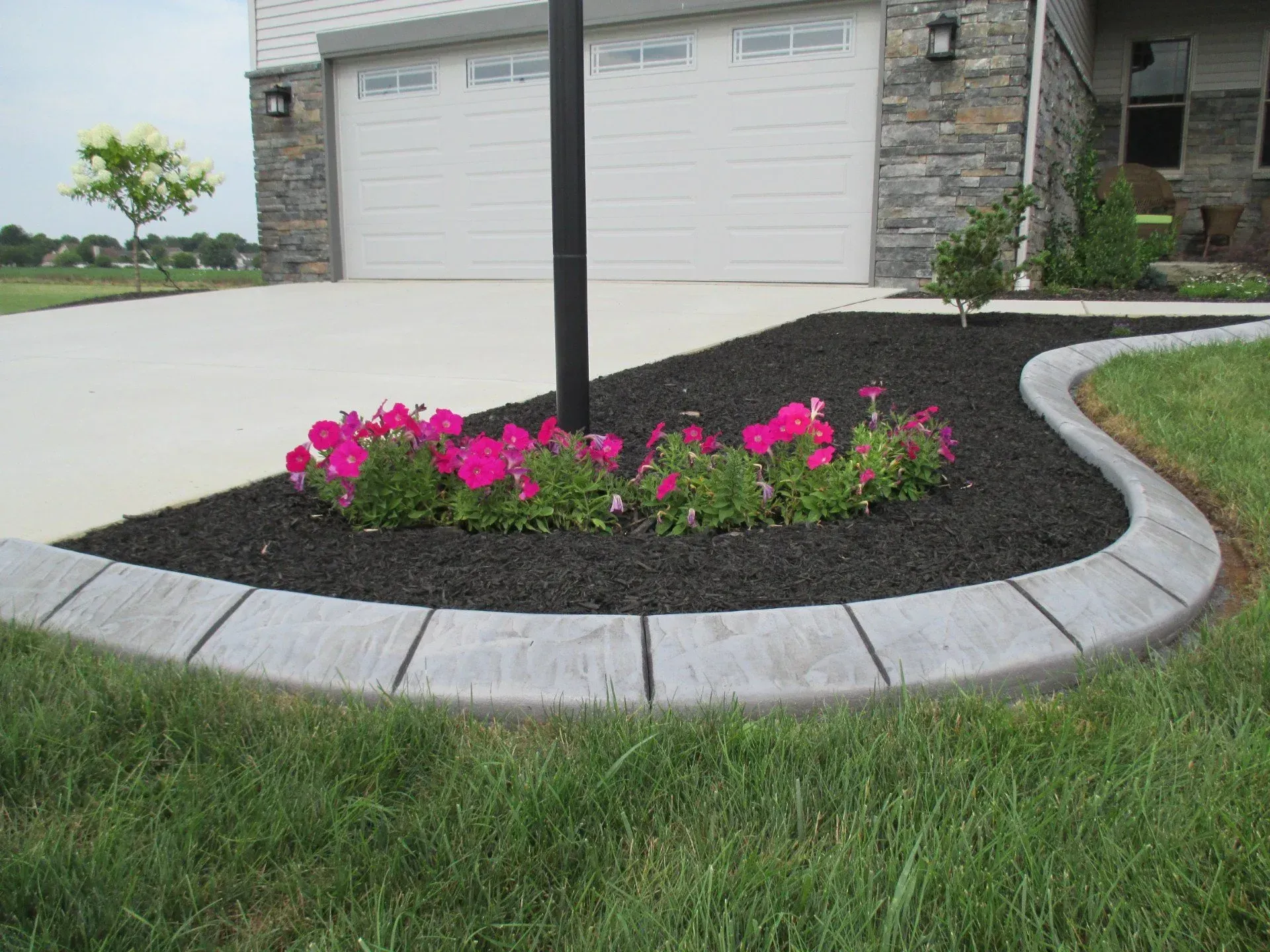 A garden bed with pink flowers and dark mulch, bordered by stone edging next to a concrete driveway and residential home.