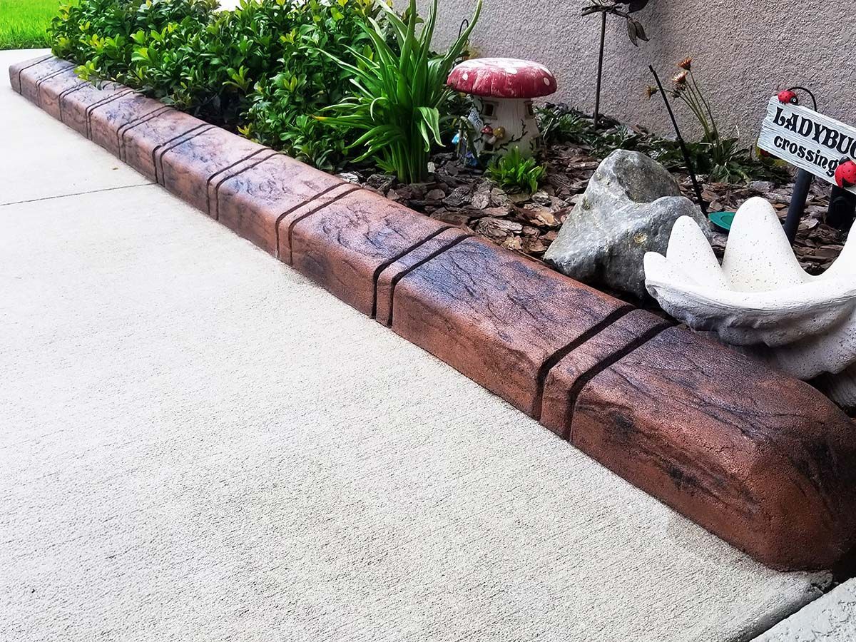A decorative concrete landscape border with a faux wood grain finish lines a flower bed next to a paved walkway.