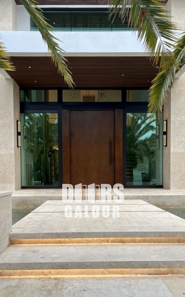 Large wooden pivot door with multipoint locking system; glass windows, palm tree visible.