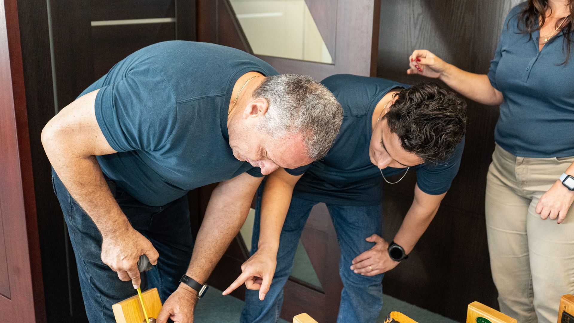 Three people bending over to inspect something on a floor with yellow tools nearby.