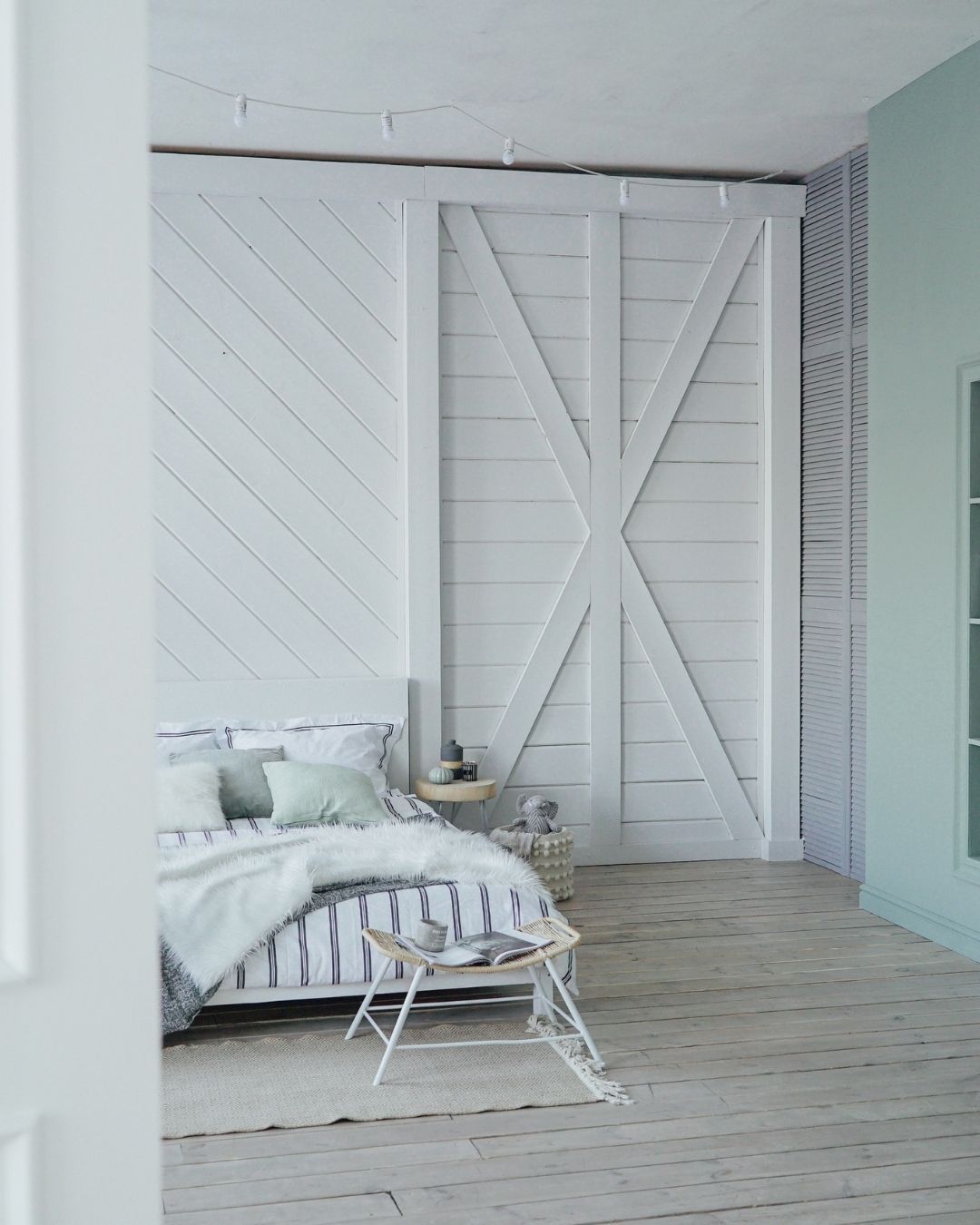 A bedroom with white wooden walls, featuring a bed with striped bedding, a white blanket, and a small metal side table.