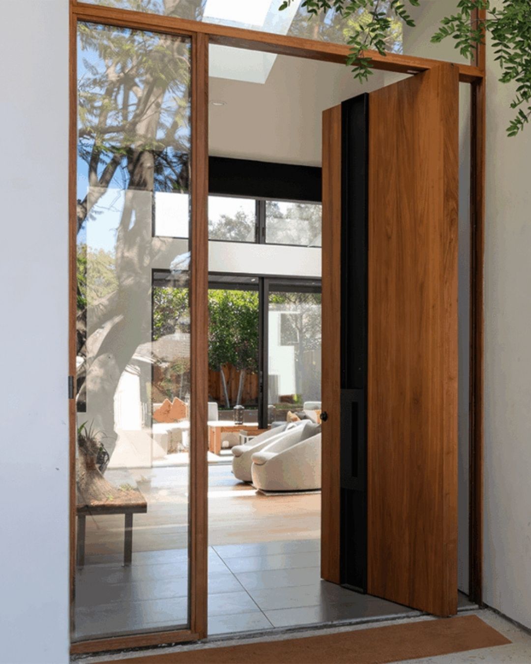 Large wooden pivot door with multipoint locking system; glass windows, palm tree visible.