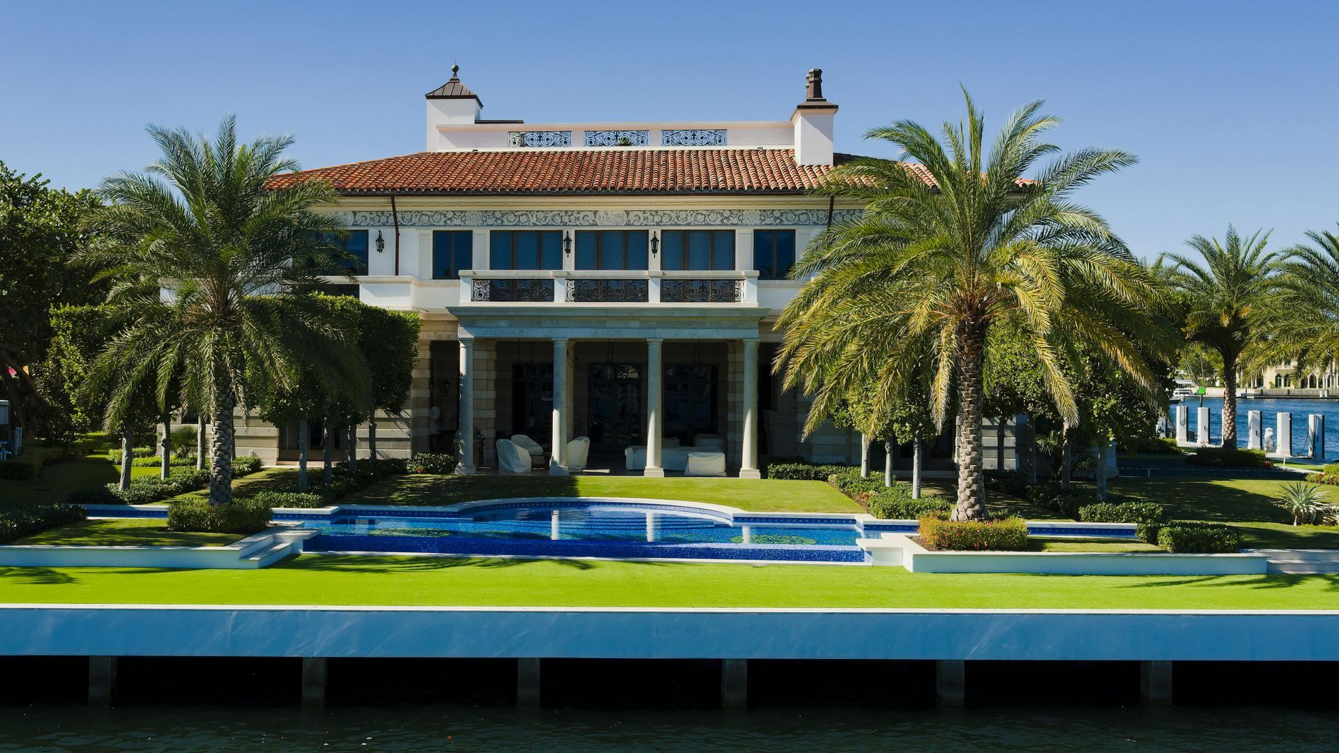 Grand villa with palm trees, a turquoise pool, and manicured lawn in bright sunlight