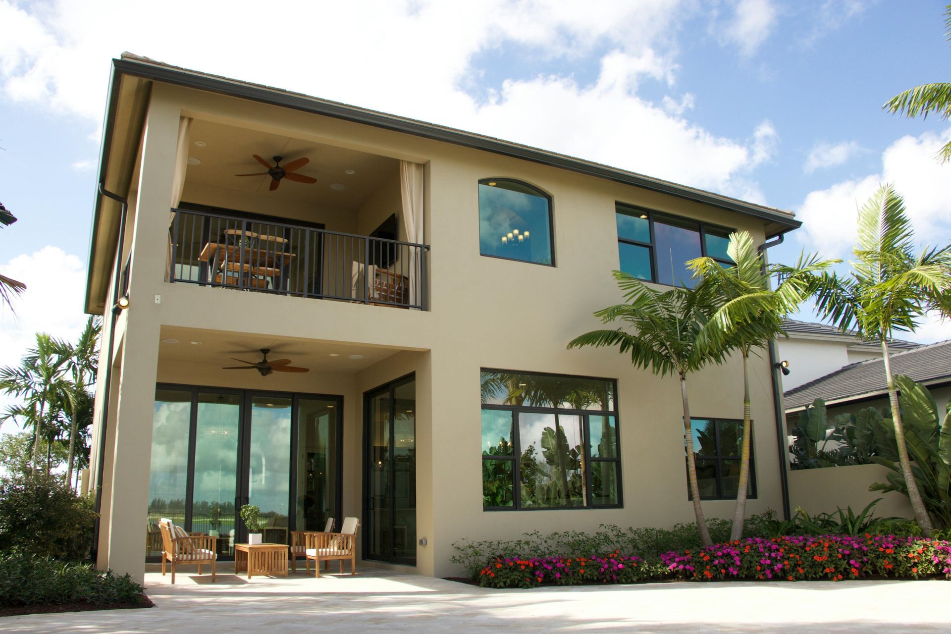 Two-story cream house with balcony, large windows, and tropical landscaping under a bright sky