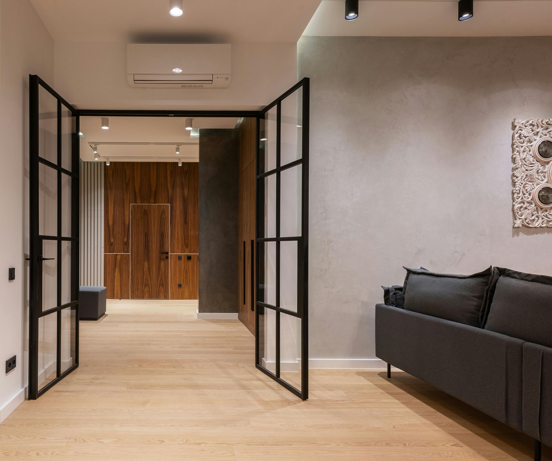 Modern room with open black-framed double doors, light wood floor, gray walls, and a dark sofa.