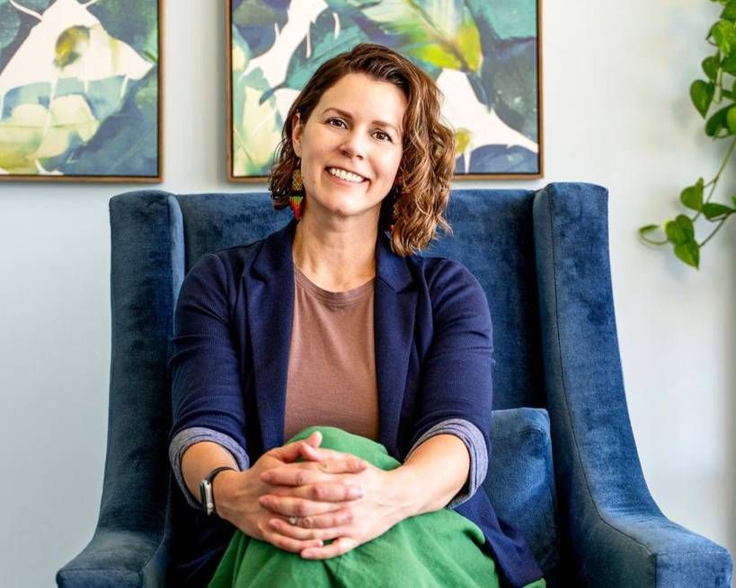 Smiling person seated in a blue armchair in a bright room with framed artwork and a leafy plant.