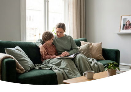 Two people sit on a green couch, looking at a tablet under a gray blanket, with a mug and plant on the table.