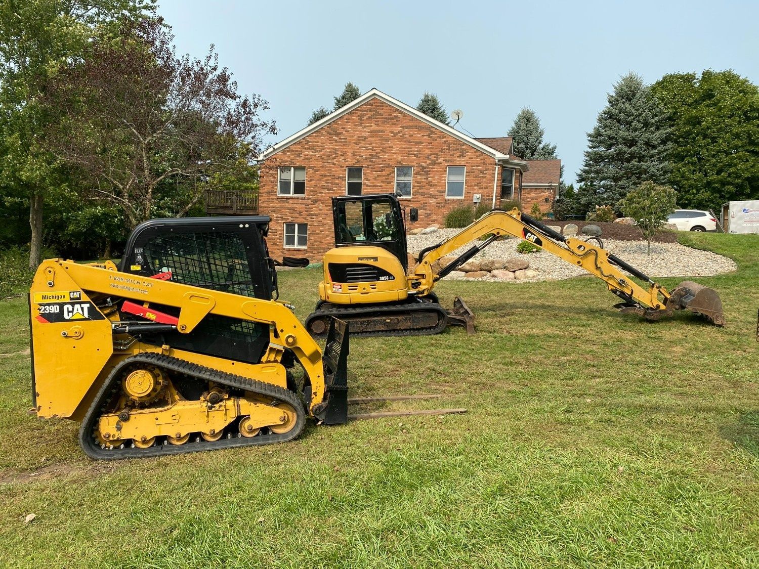 Yellow and black mini skid steer in a grassy yard with a brick house and tree pile in the background