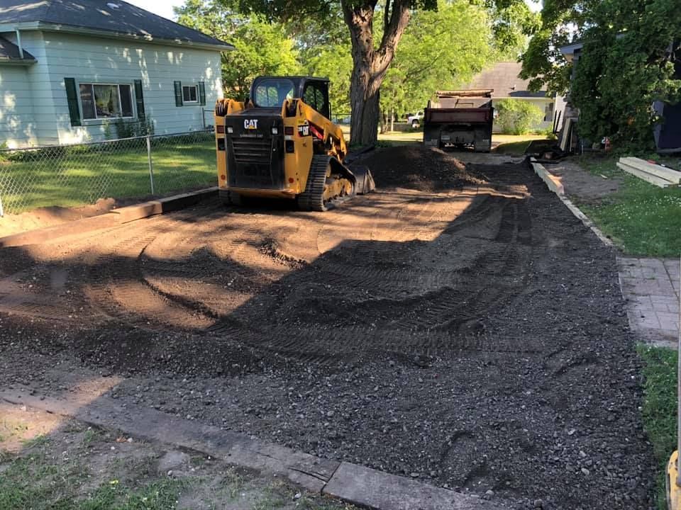 Small excavator grading a driveway beside a house, with fresh gravel and dirt piled nearby.