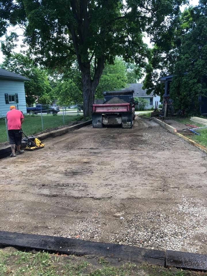 Driveway under construction with a dump truck, worker, and newly leveled gravel area under trees