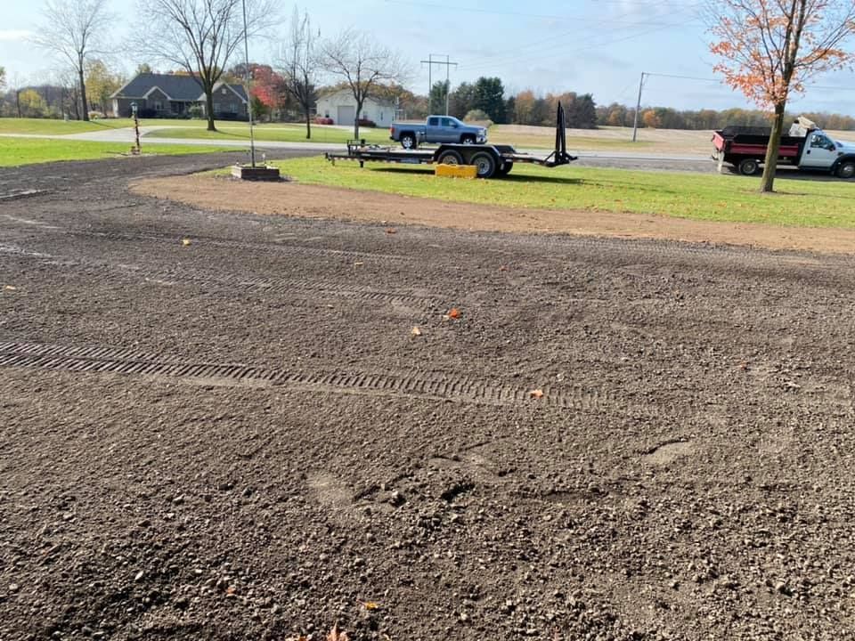 Graded dirt lot with a pickup truck, trailer, and parked cars near a road on a sunny day