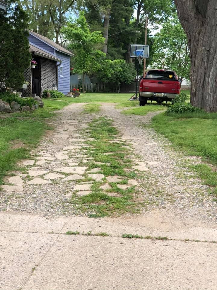 Cracked gravel driveway beside a house, with a red car parked near a basketball hoop and trees lining the yard.