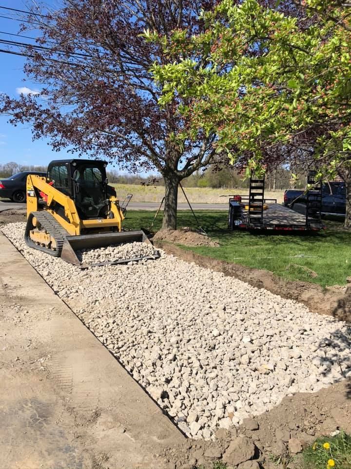 Skid steer spreading white gravel along a driveway beside a tree and parked truck