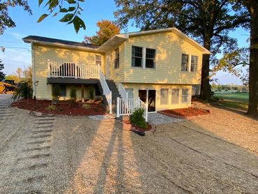 Yellow house with white trim, front porch, and gravel driveway in a wooded yard at sunset