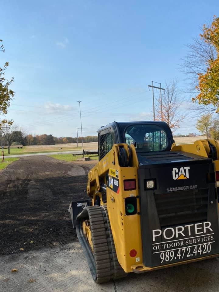 Yellow CAT skid steer with PORTER Services lettering, parked on gravel near an open field on a sunny day.