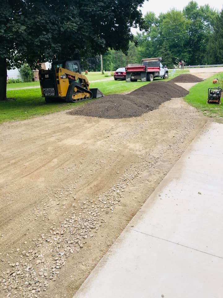 Roadwork on a sidewalk edge with a skid steer, gravel pile, and red pickup truck nearby.