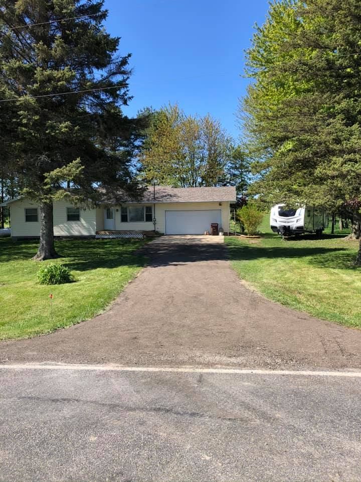 Single-story house with a long driveway, green lawn, trees, and a camper parked on the right.