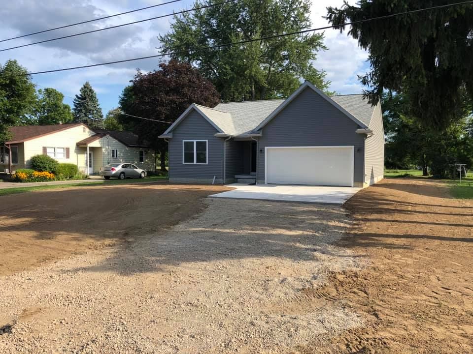 Gray house with white garage on a gravel driveway, surrounded by trees under a cloudy sky