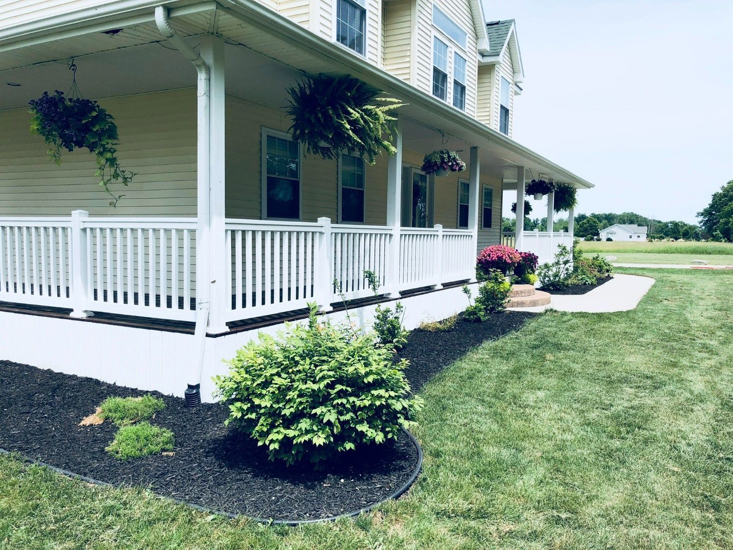 White house porch with railings, hanging plants, and a landscaped lawn on a sunny day