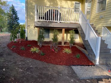 Landscaped front porch with white railings, stairs, and red mulch beds beside a yellow house