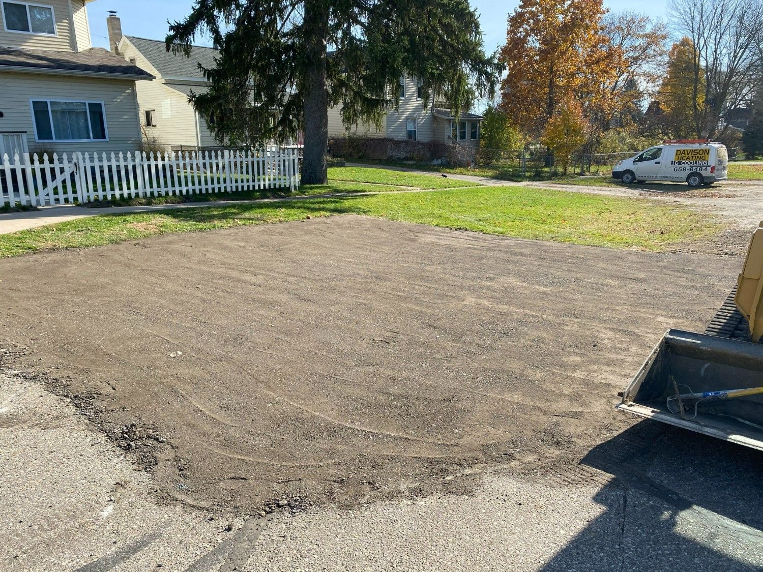 Gravel driveway beside a white house and picket fence, with a tree and parked work van in the background.