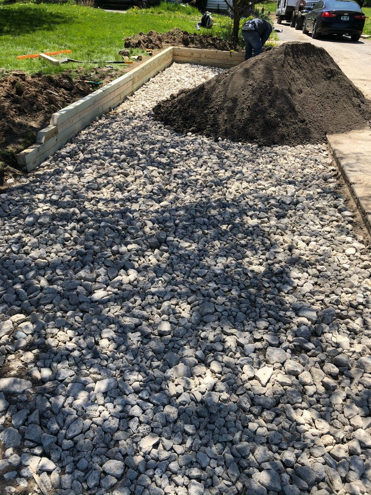 Fresh gravel driveway beside a curb, with a pile of dark gravel and a parked car in the background