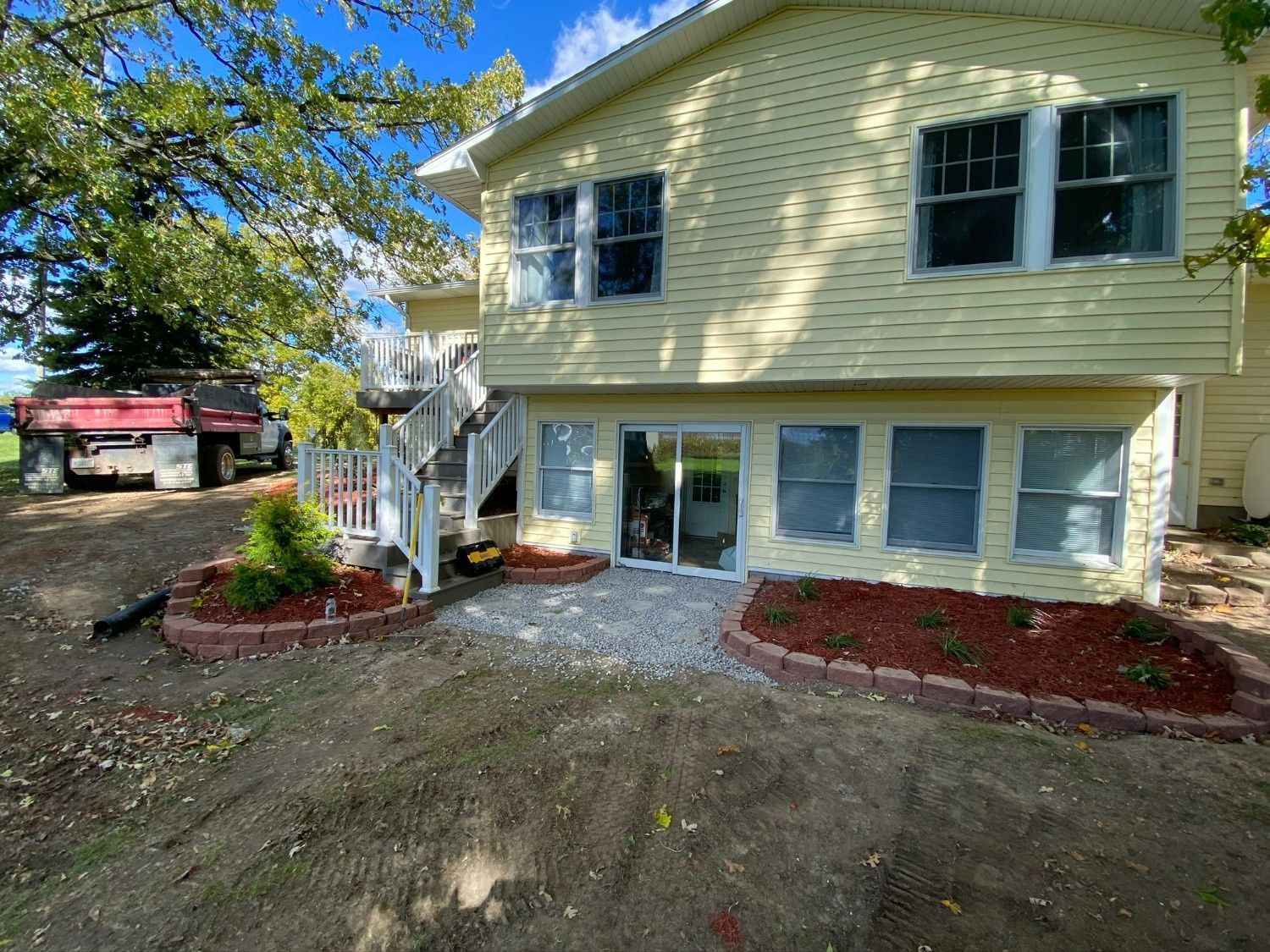 Yellow two-story house with basement windows, white stairs, gravel path, and landscaped front beds