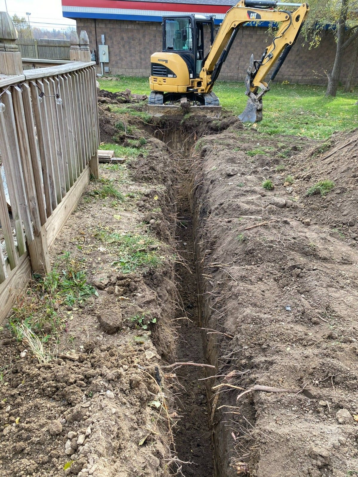 Backhoe digging a narrow trench beside a wooden fence in a muddy yard