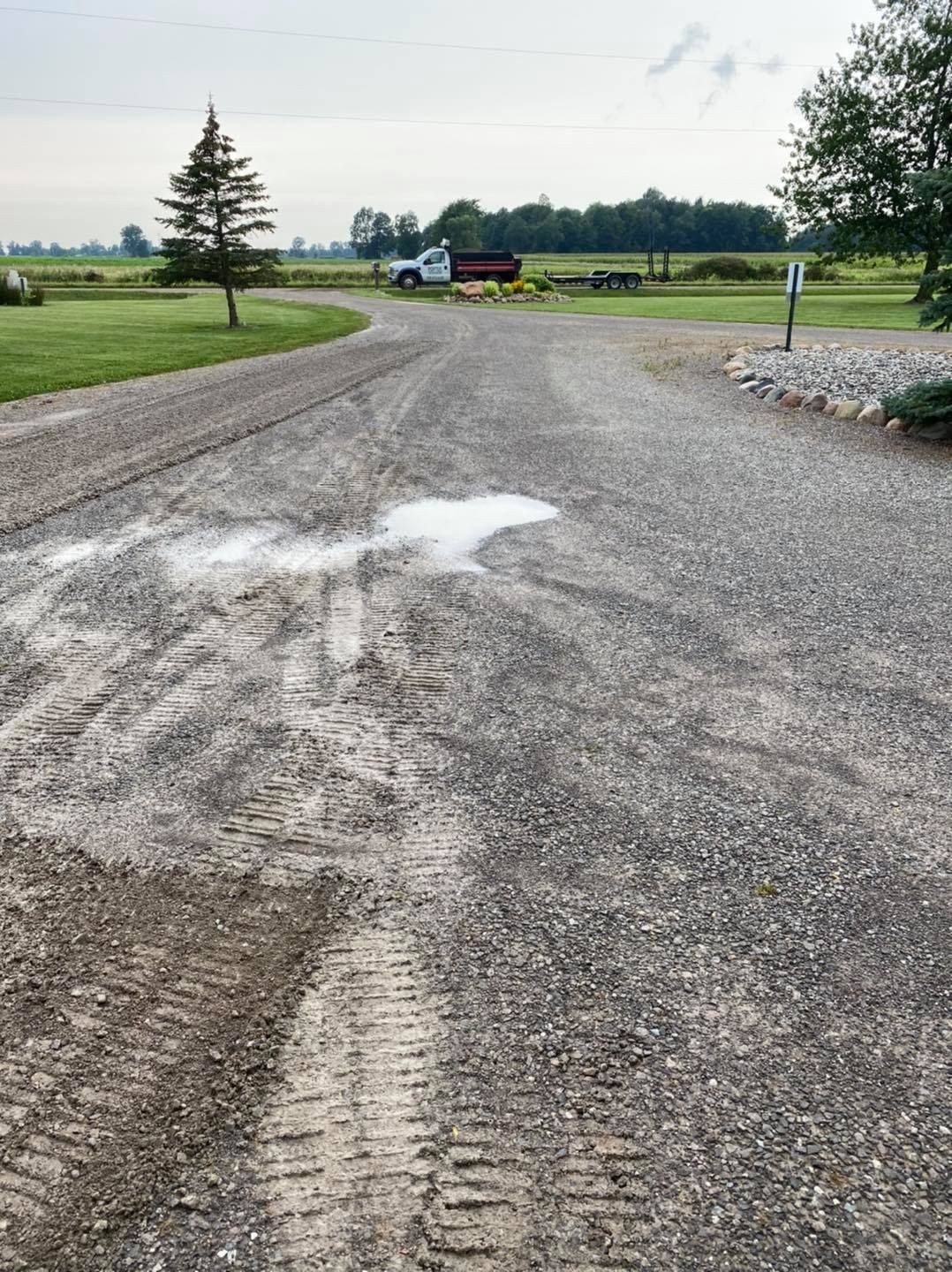 Gravel driveway with tire tracks leading to a grassy rural road and a parked farm trailer in the distance
