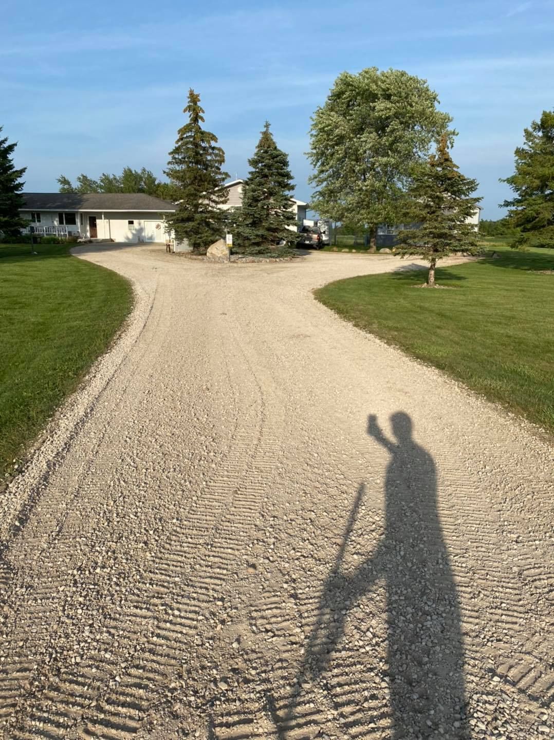 Gravel driveway leading to a house and trees on a sunny day, with a long shadow in the foreground