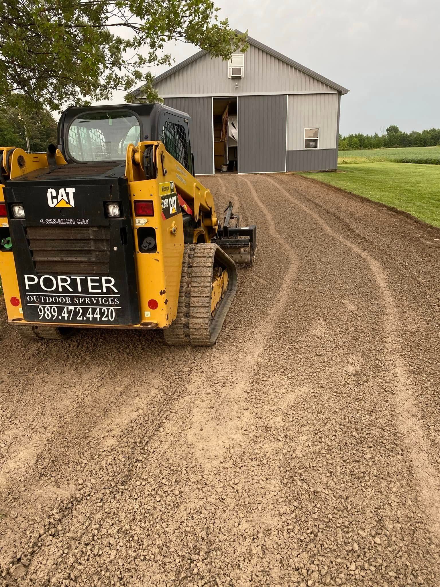 Yellow skid steer on gravel driveway beside a barn and grassy field under overcast sky