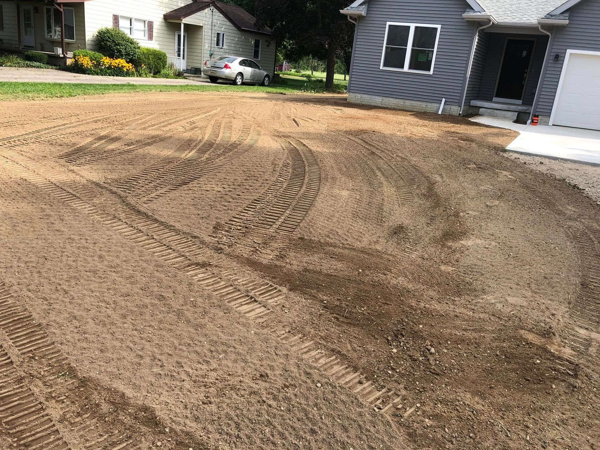 Freshly graded dirt driveway beside a house, with tire tracks and a parked vehicle in the background