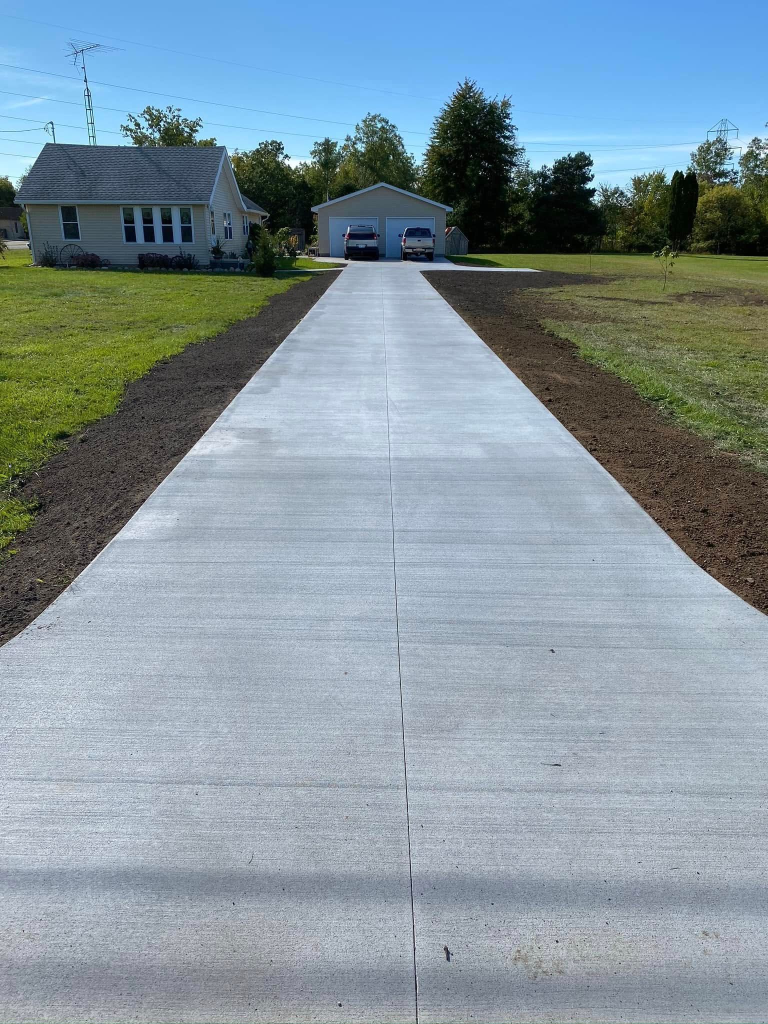 Concrete driveway leading to a house and detached garage on a sunny rural property.