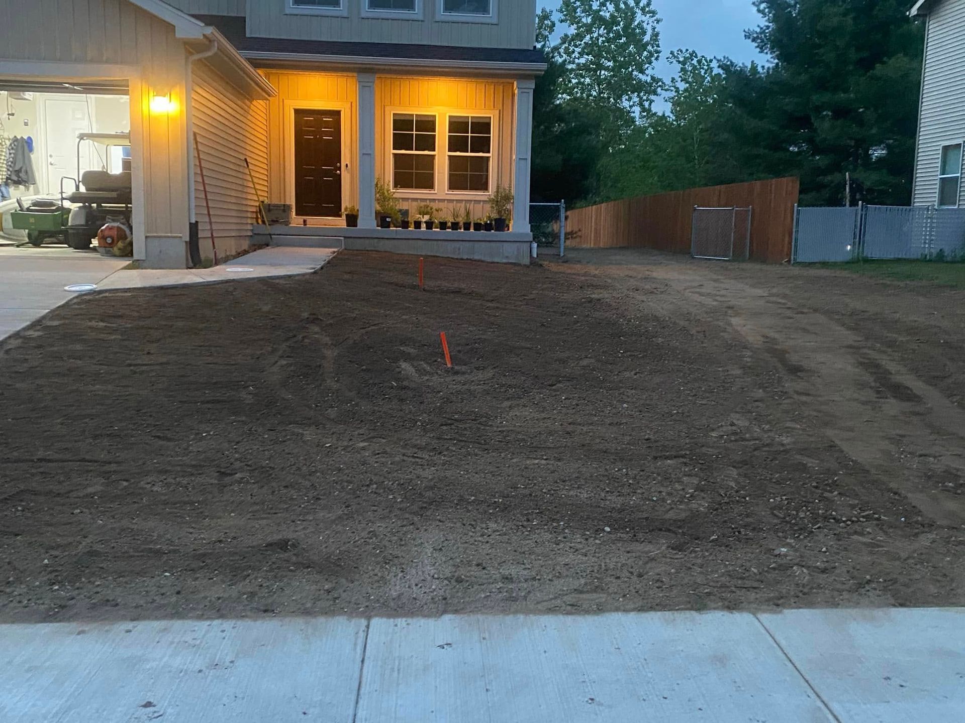 Freshly graded front yard beside a house at dusk, with lit porch and windows.