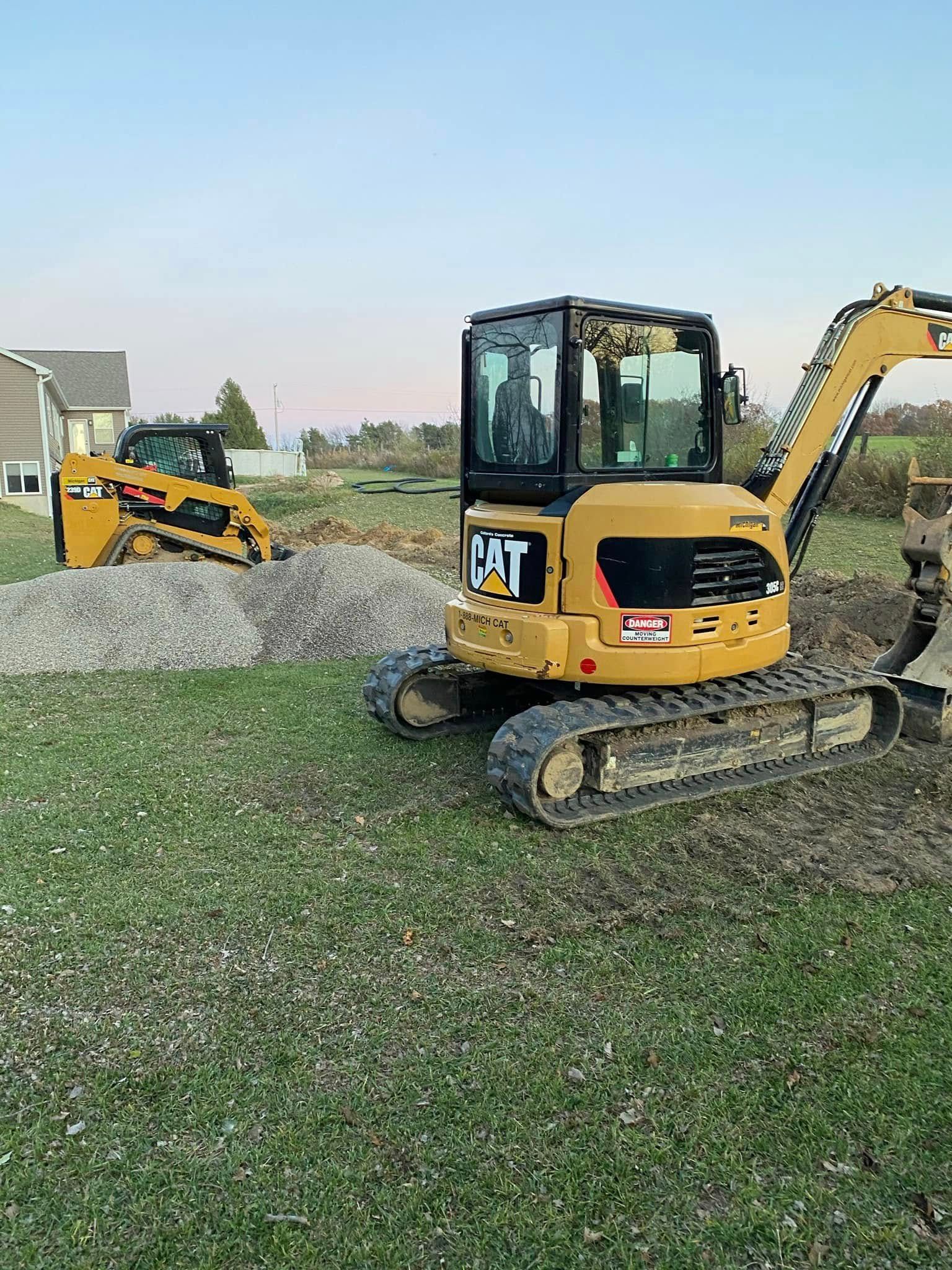 Yellow CAT excavator on a gravel pile beside another construction machine in a grassy field at dusk