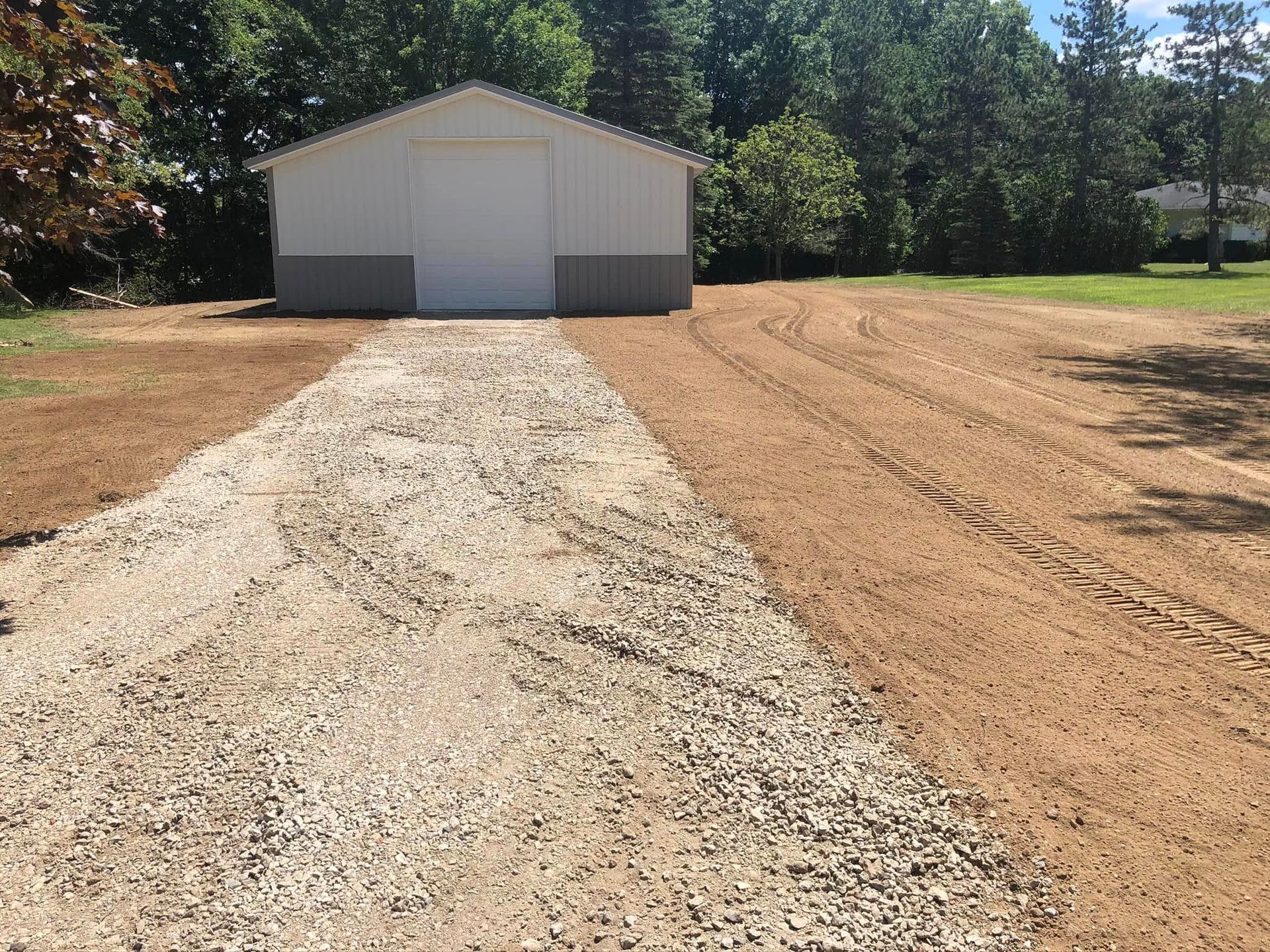 Gravel driveway leading to a white garage with a dirt lot and trees in the background